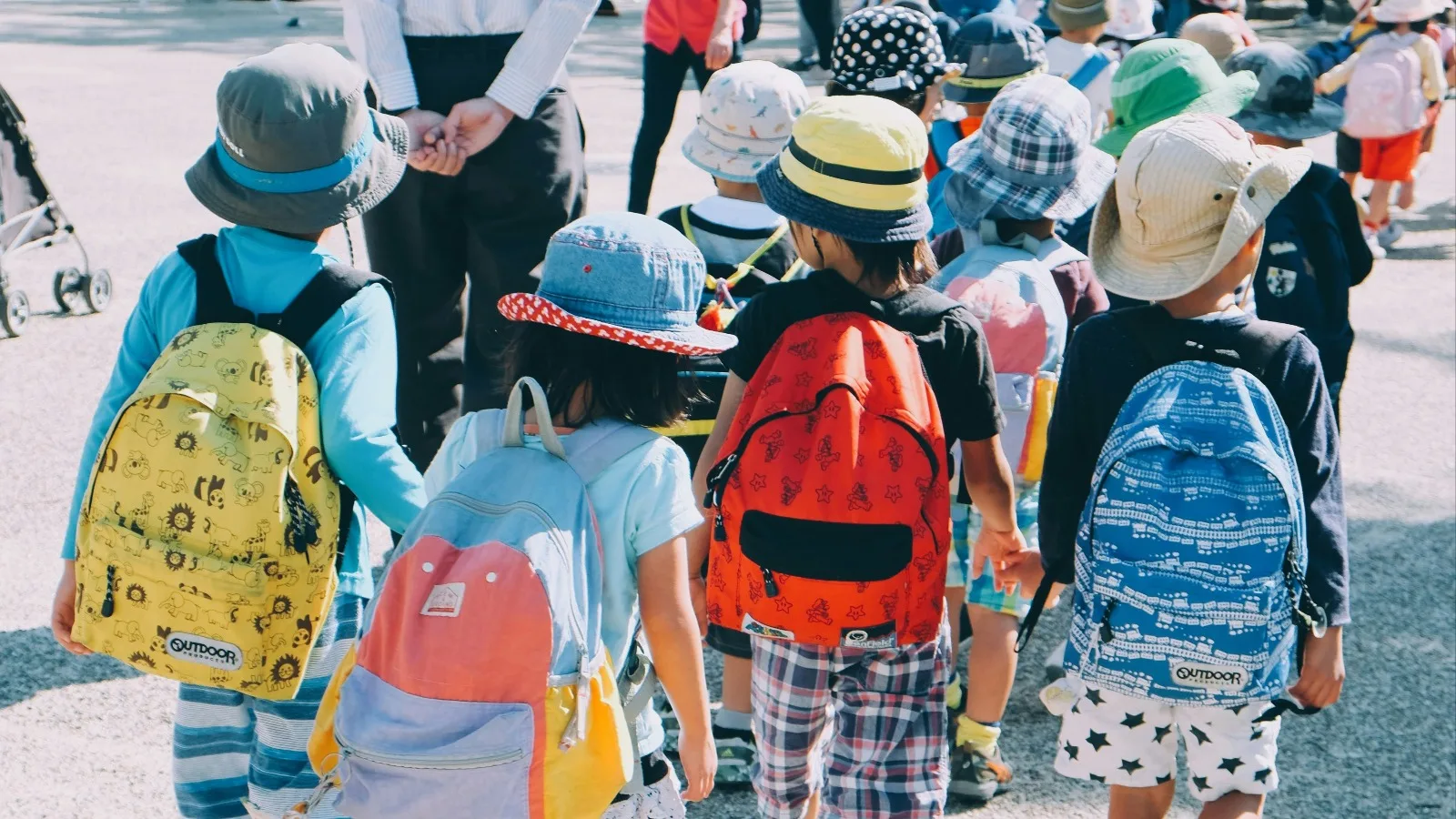 Un groupe d'enfants coiffés de chapeaux et portant des sacs à dos colorés, vus de dos, marchant en rang