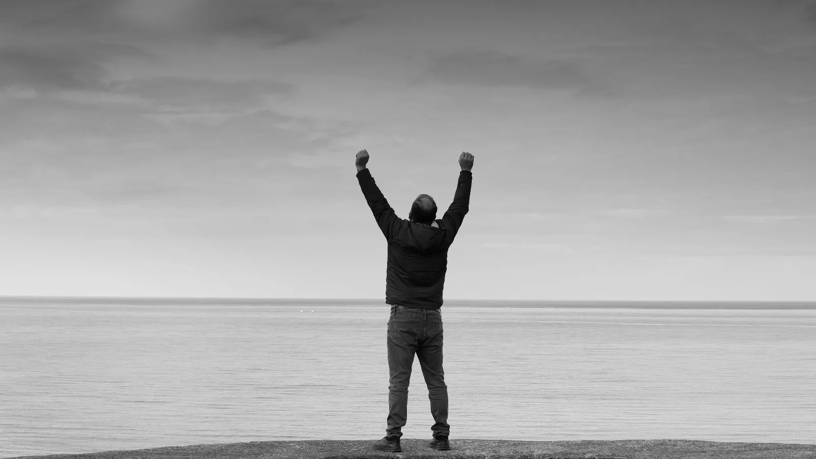 Un homme debout face à la mer, bras levés vers le ciel, photographié en noir et blanc