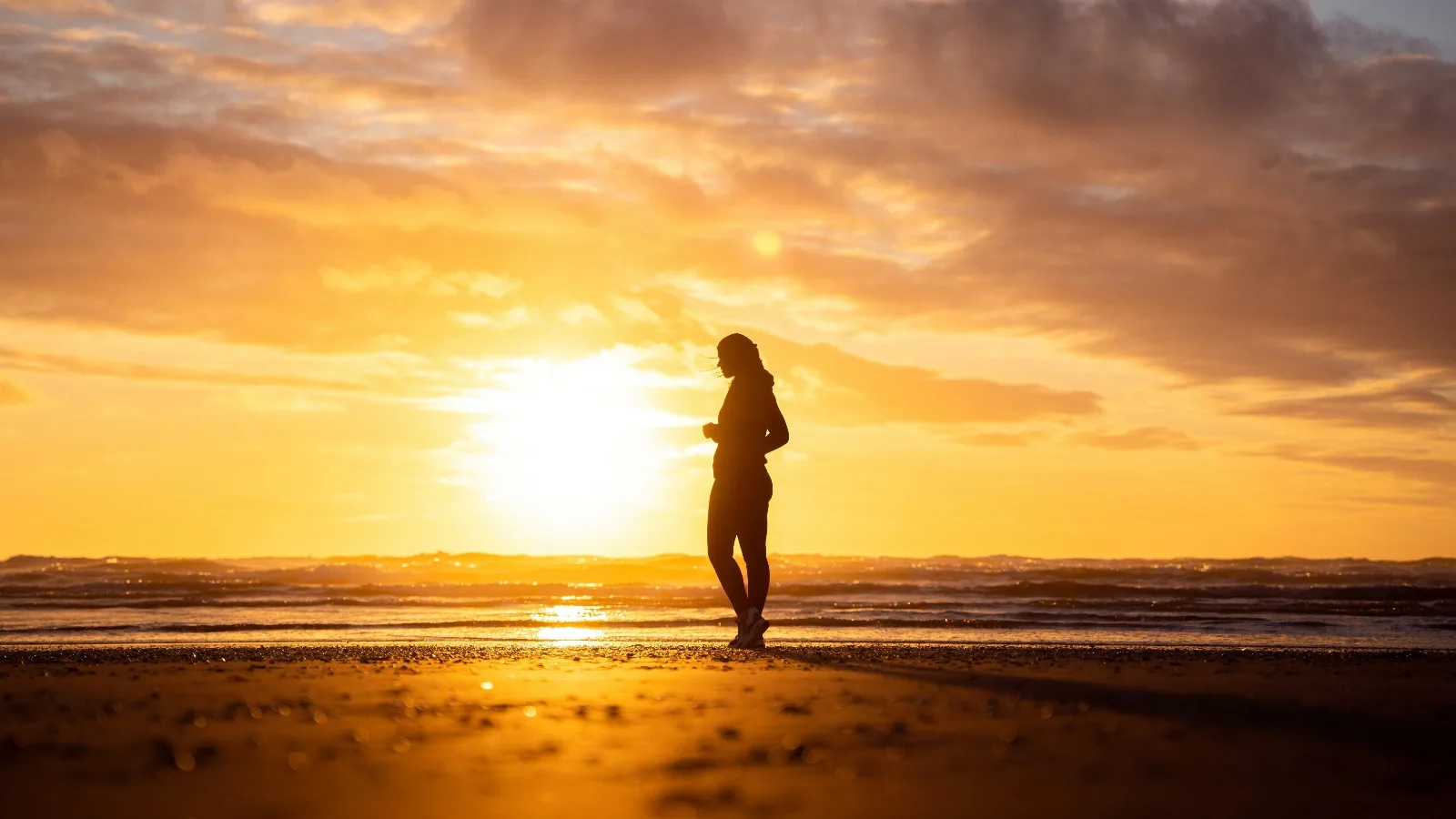 La silhouette d'une personne marchant sur une plage à contre-jour, face au soleil couchant